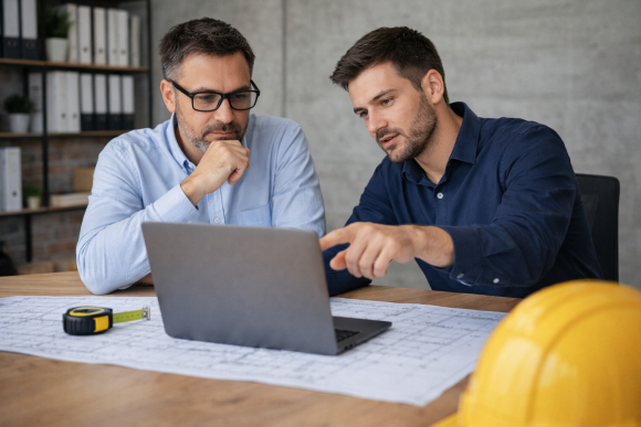 Zwei Bauprofis arbeiten in einem Büro an ASBwin auf dem Laptop – das Arrangement symbolisiert düberzeugende Gründe für die AVA-Software.