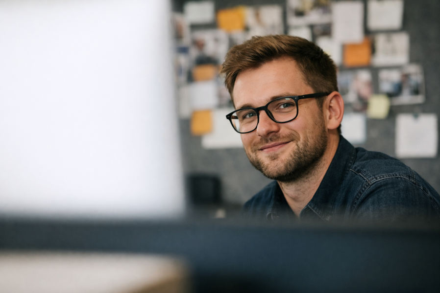Mann arbeitet konzentriert im Büro am Computer mit Leistungsverzeichnis-Nummerierung, auf dem Schreibtisch liegen Baupläne, Notizbuch und gelber Bauhelm