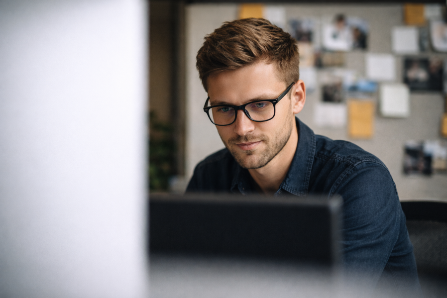 Mann arbeitet konzentriert im Büro am Laptop, auf dem Schreibtisch liegen ein Notizbuch, ein Stift und ein gelber Bauhelm